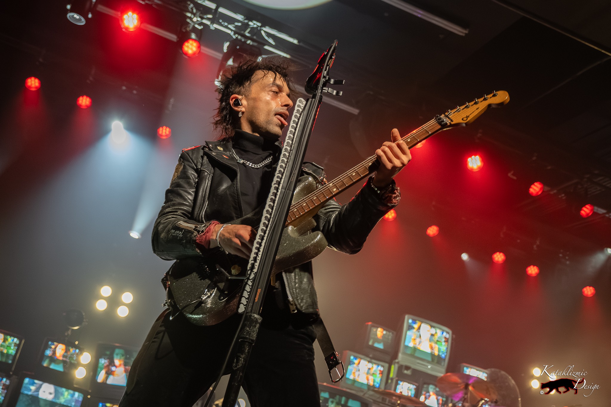 BOYS LIKE GIRLS guitarist Jamel Hawke performs under red stage lights with vintage televisions behind him.