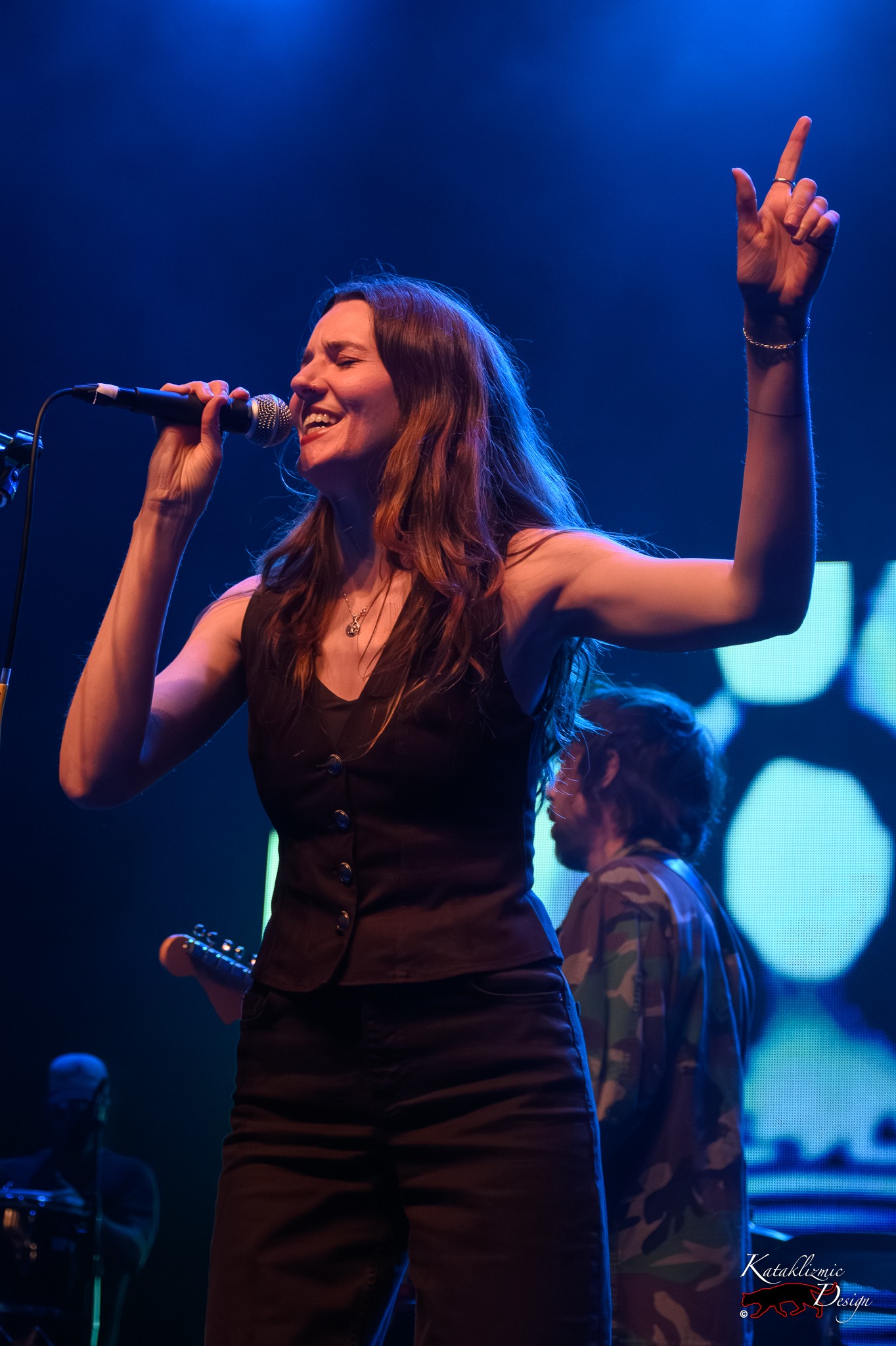 Jill Harris sings with Broken Social Scene, raising one arm under blue stage lights.