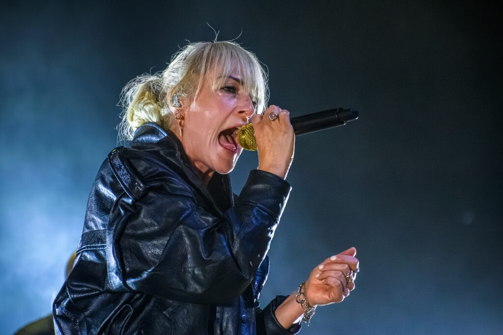 Emily Haines of Metric sings into a gold microphone under blue stage lighting.
