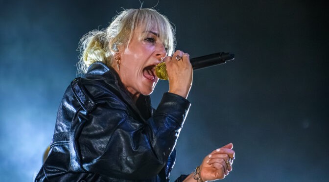 Emily Haines of Metric sings into a gold microphone under blue stage lighting.
