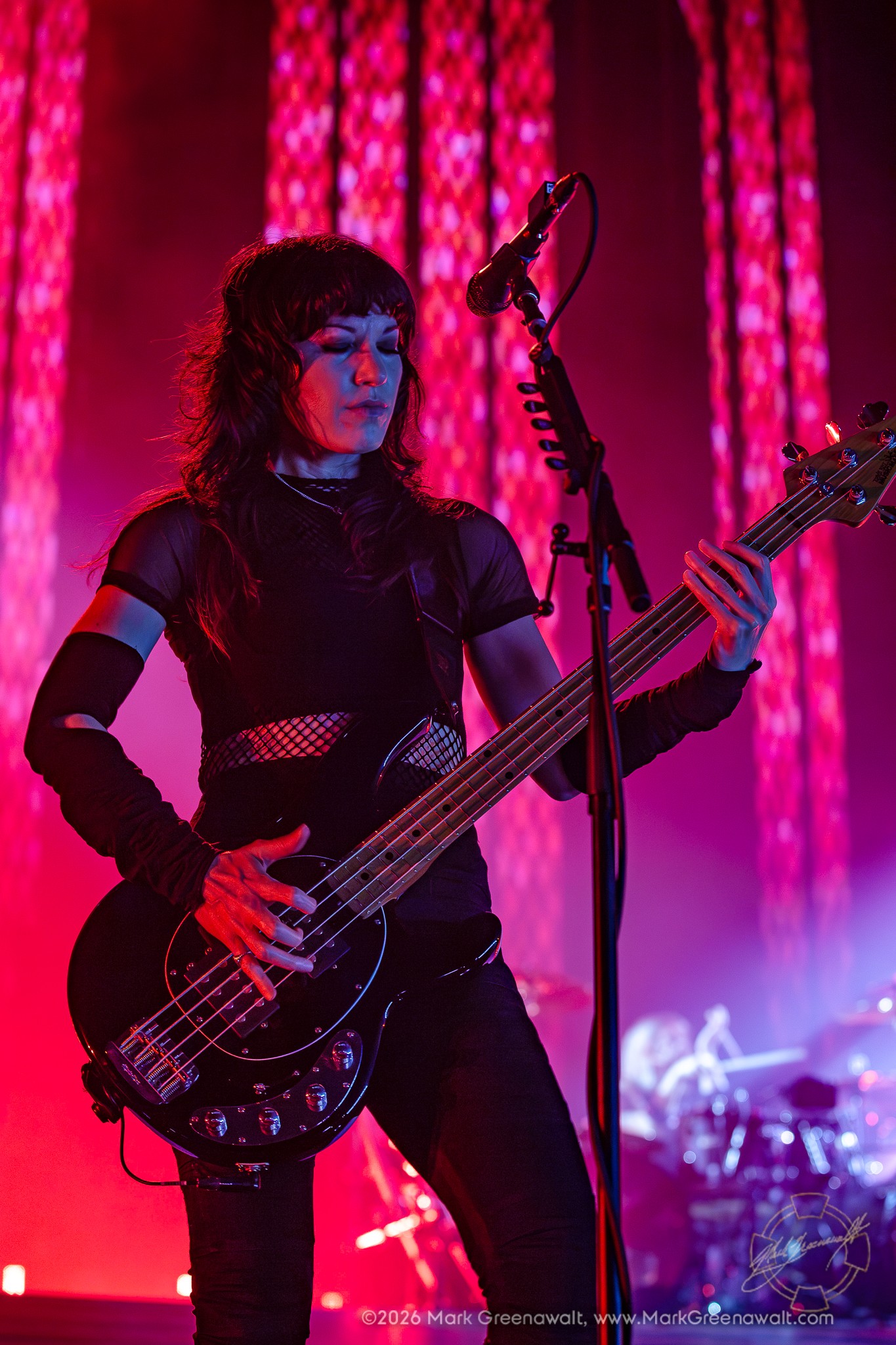 Emma Anzai plays a black bass beneath vivid red and magenta lighting, with tall cathedral-inspired arches glowing behind her and the drummer blurred in the distance.