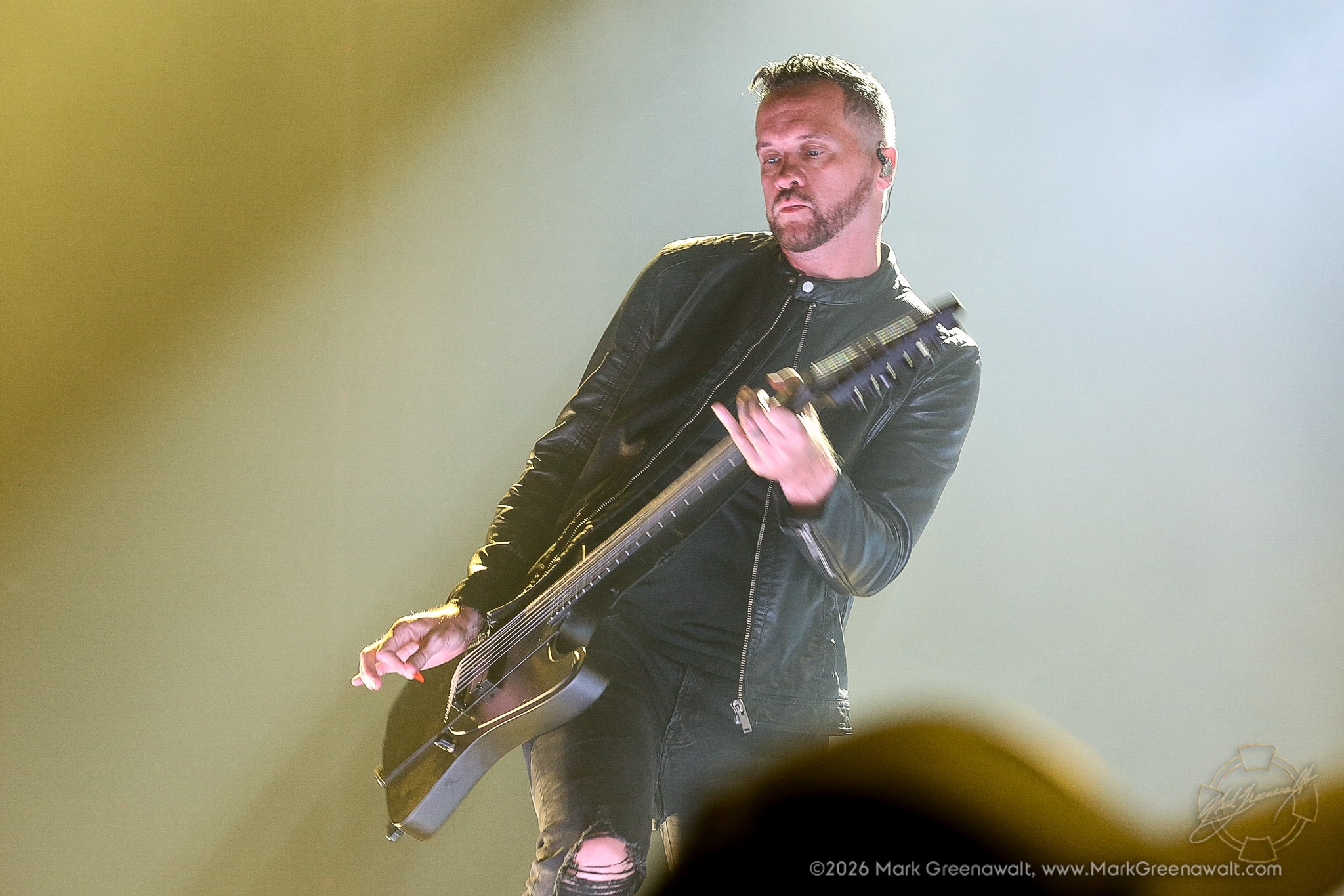 Troy McLawhorn plays an angular black electric guitar while leaning to one side in a black leather jacket and ripped jeans, illuminated by bright white and golden stage light.