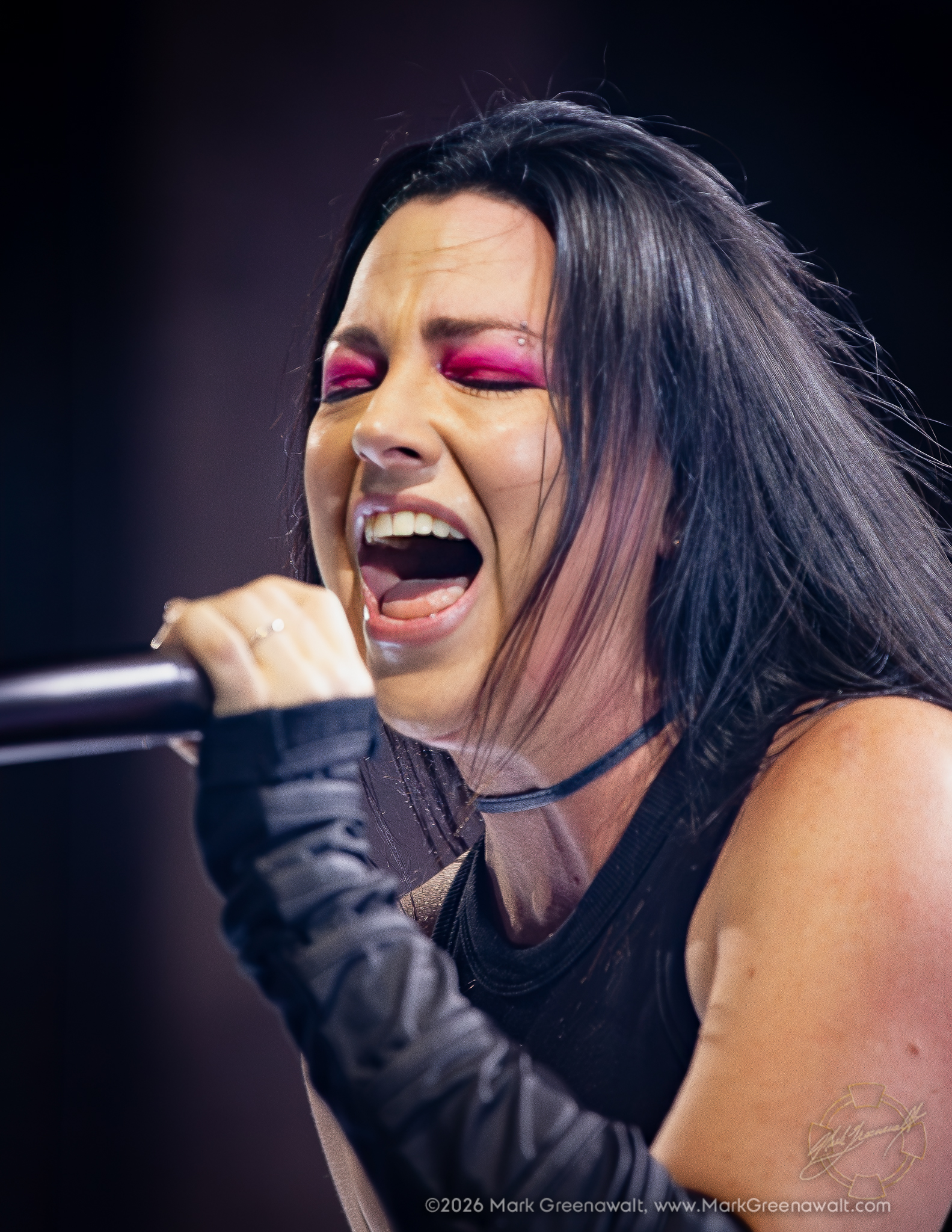 Tight close-up of Amy Lee singing with her eyes closed and mouth open, vivid magenta eye makeup contrasting with her dark hair and black stagewear.