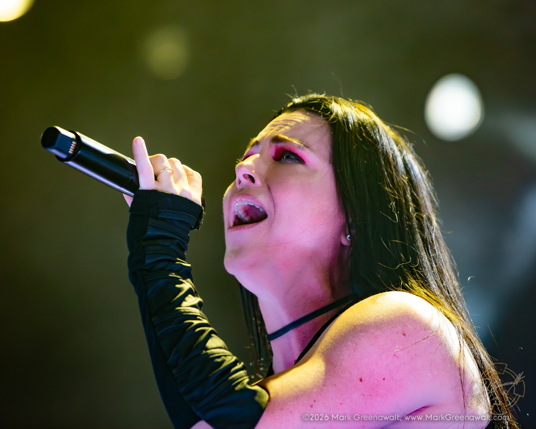 Tight three-quarter close-up of Amy Lee singing into a black handheld microphone, her vivid pink eye makeup and long dark hair illuminated by warm yellow-green stage lights.