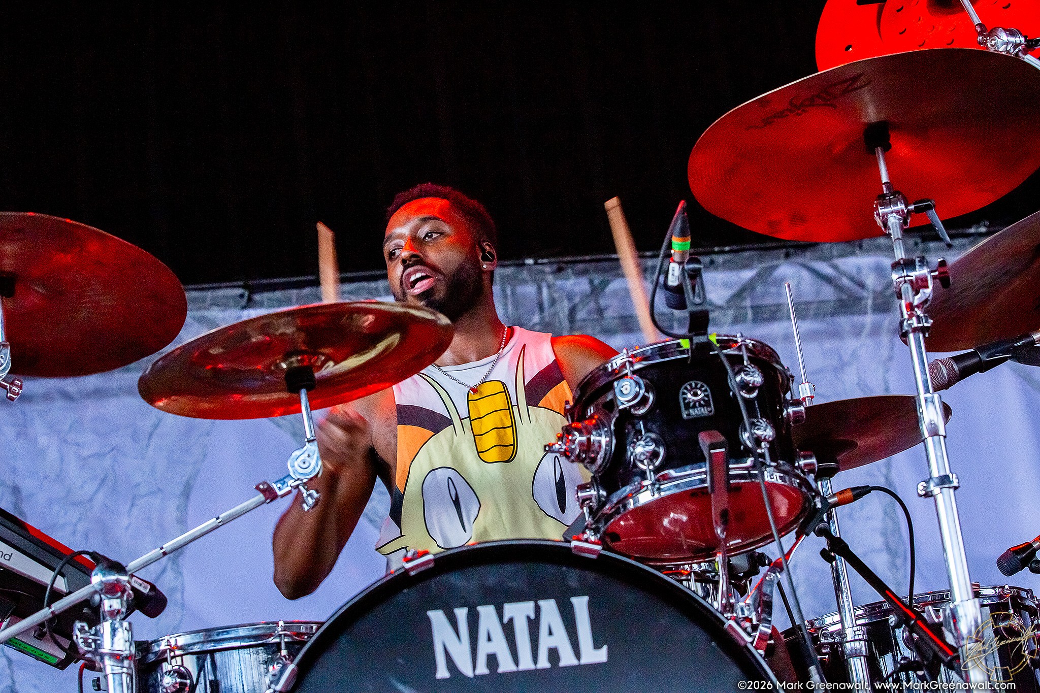 Nova Twins touring drummer Jake Woodward performs behind a large drum kit, raising both drumsticks mid-strike beneath red stage lighting.