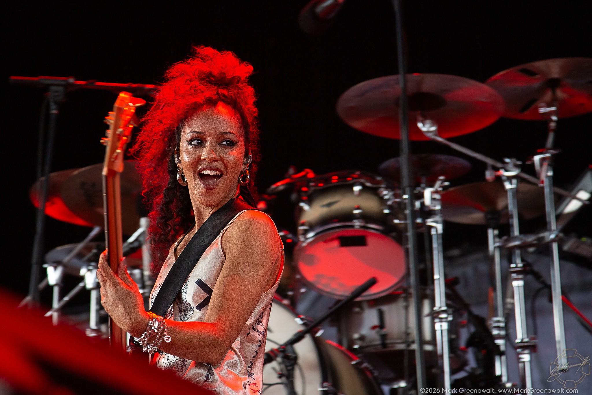 Nova Twins bassist Georgia South performs with an animated, open-mouthed expression while holding her bass upright under red stage lighting, with a drum kit behind her
