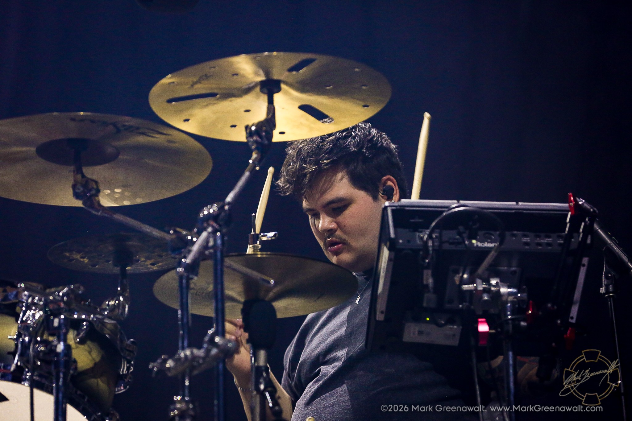 Zev Rosenberg sits behind an expansive drum kit, looking downward as cymbals surround him and an electronic percussion pad partially obscures his right side.