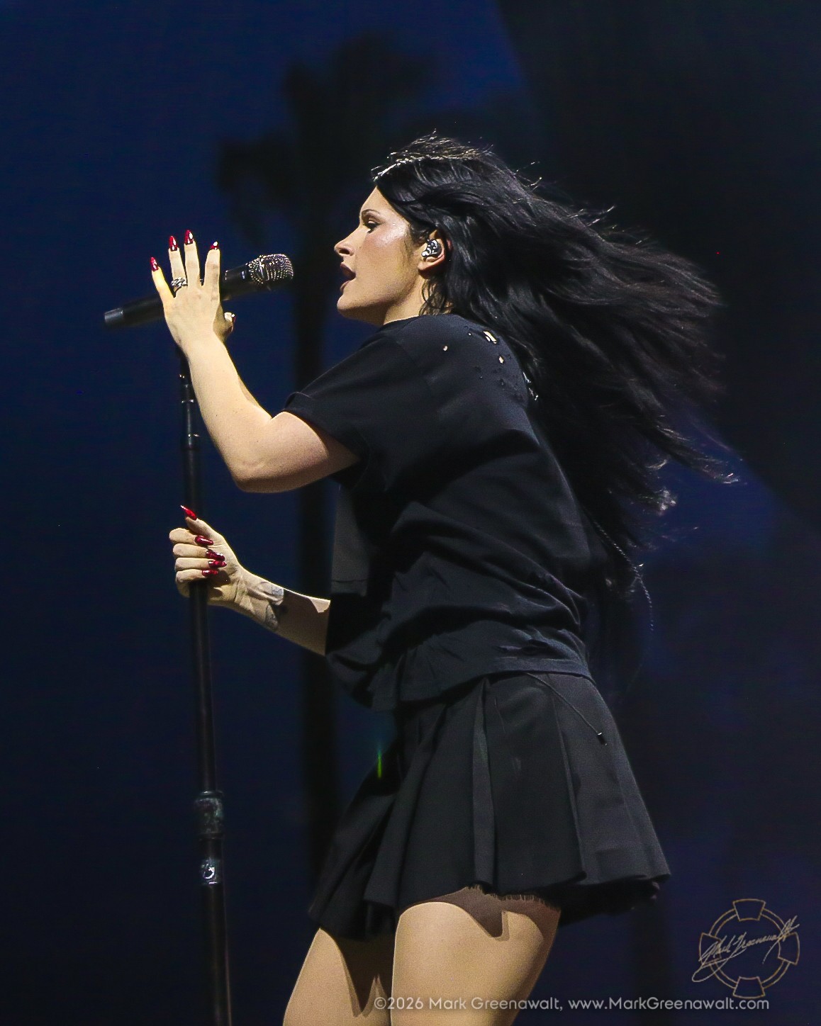 Side-profile view of Courtney LaPlante singing at a microphone stand in an all-black outfit, with her long dark hair streaming dramatically behind her and red nails visible against the dim stage.