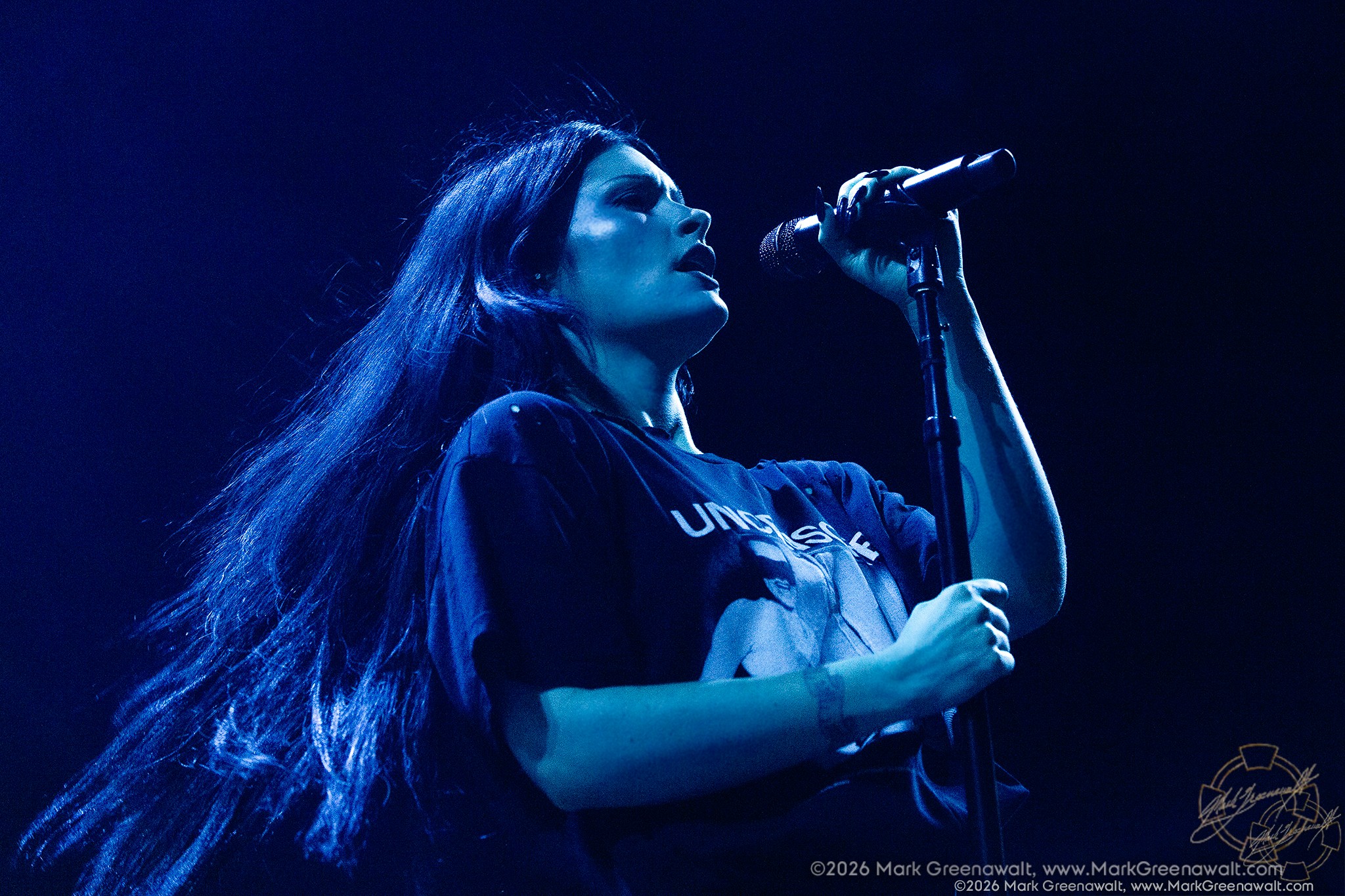 Low-angle close-up of Courtney LaPlante singing into a microphone beneath deep blue lighting, one hand gripping the mic and the other resting on the stand as her long dark hair flows behind her