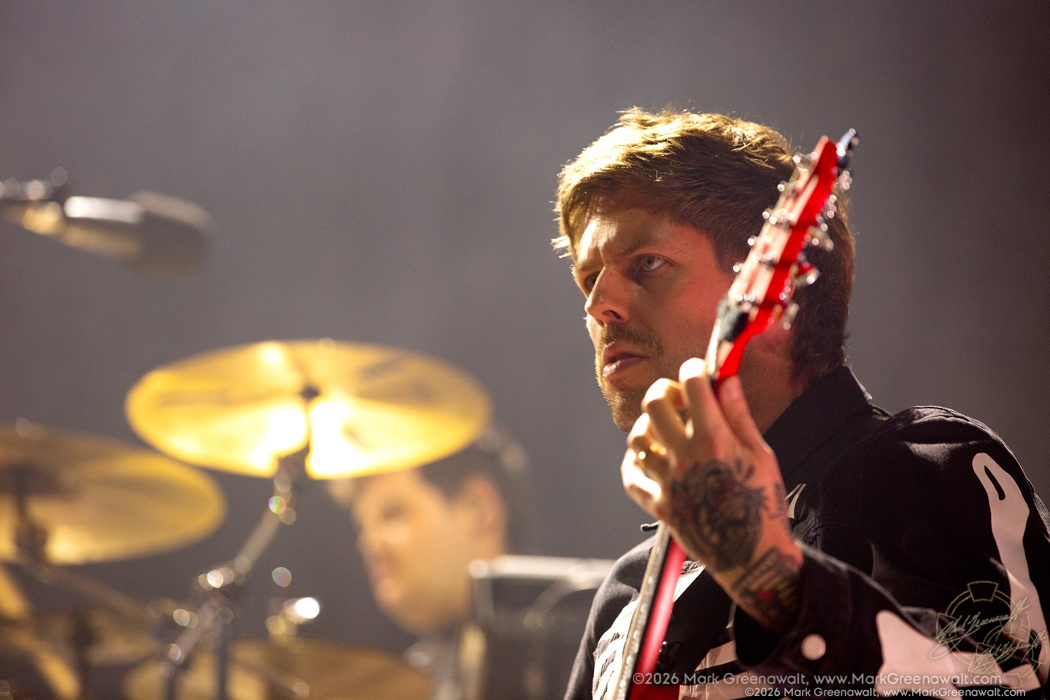 Close-up of Mike Stringer playing a red guitar, his tattooed hand gripping the neck as warm stage light illuminates his face and a drummer appears softly blurred behind him.