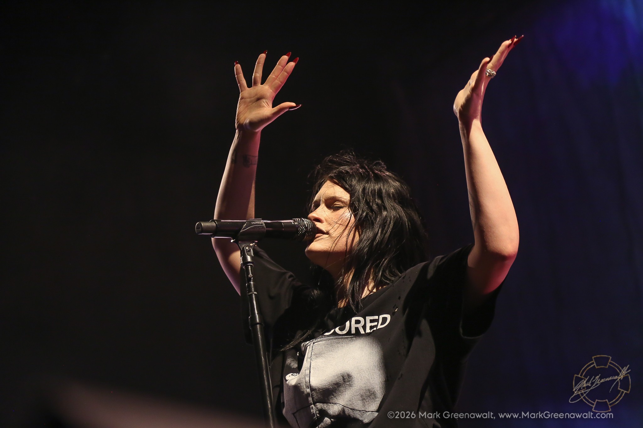 Courtney LaPlante stands at a microphone with both hands lifted high above her head and fingers spread, framed against a dark stage with faint purple lighting.