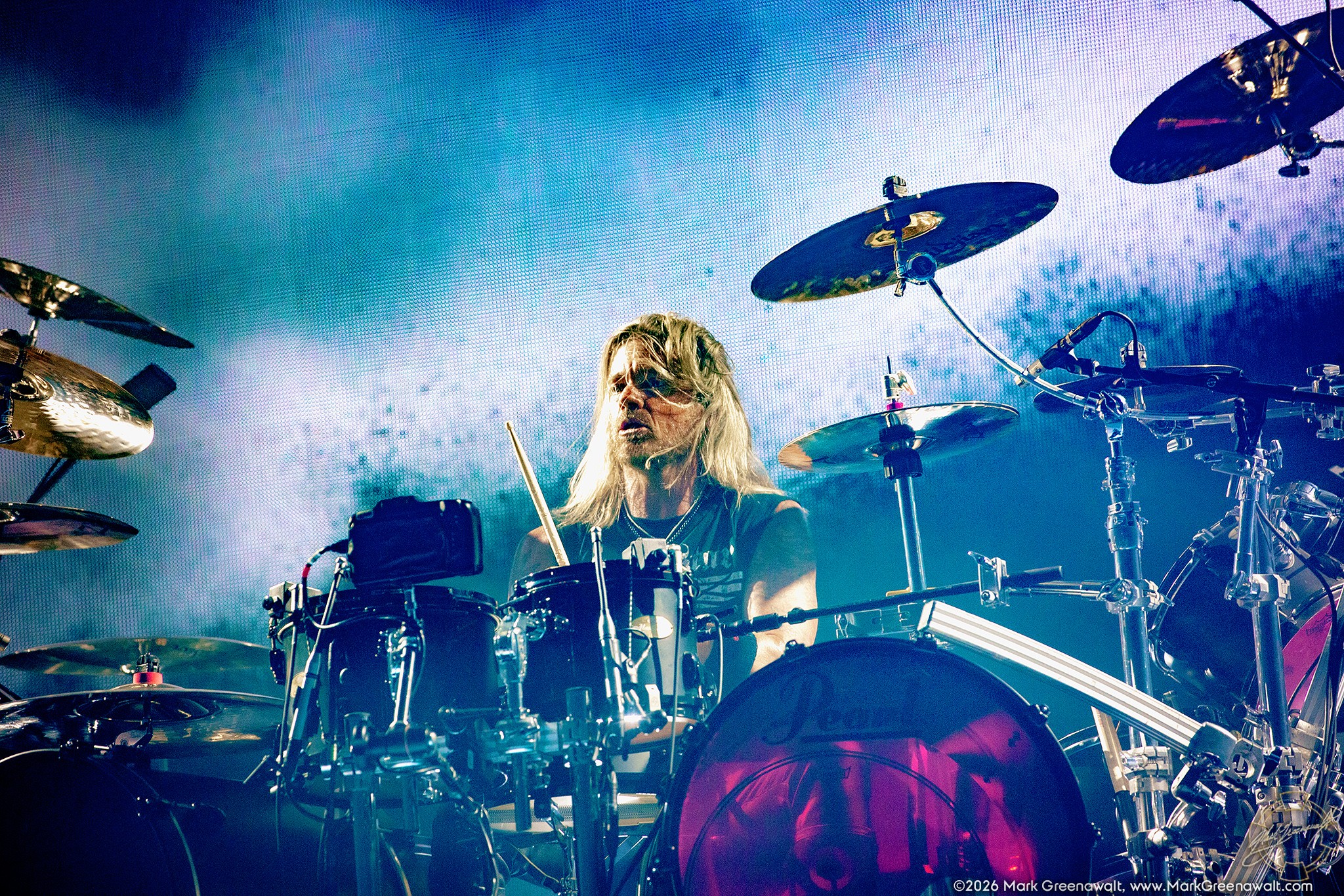 Will Hunt plays amid a large drum setup, his long blond hair falling across his face as cymbals frame him against a bright blue-and-white video backdrop.