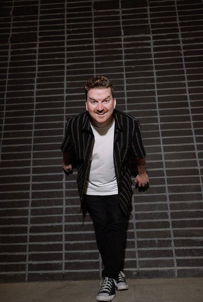 Phoenix pop-punk artist Mike Goodrick smiling in front of a dark brick wall, wearing a black striped shirt, white T-shirt, black pants, and sneakers.