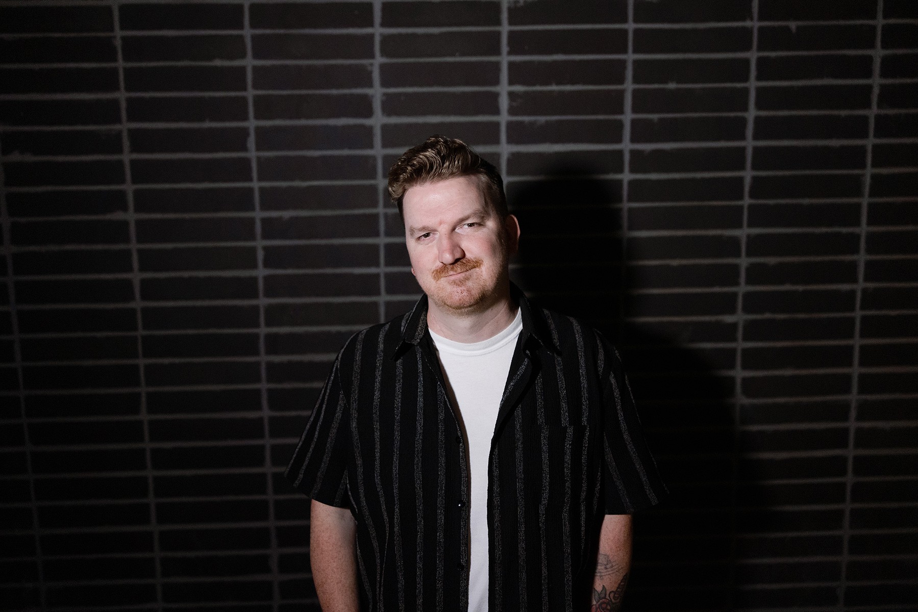 Phoenix pop-punk artist Mike Goodrick posing in front of a dark brick wall, wearing a black striped shirt over a white T-shirt.
