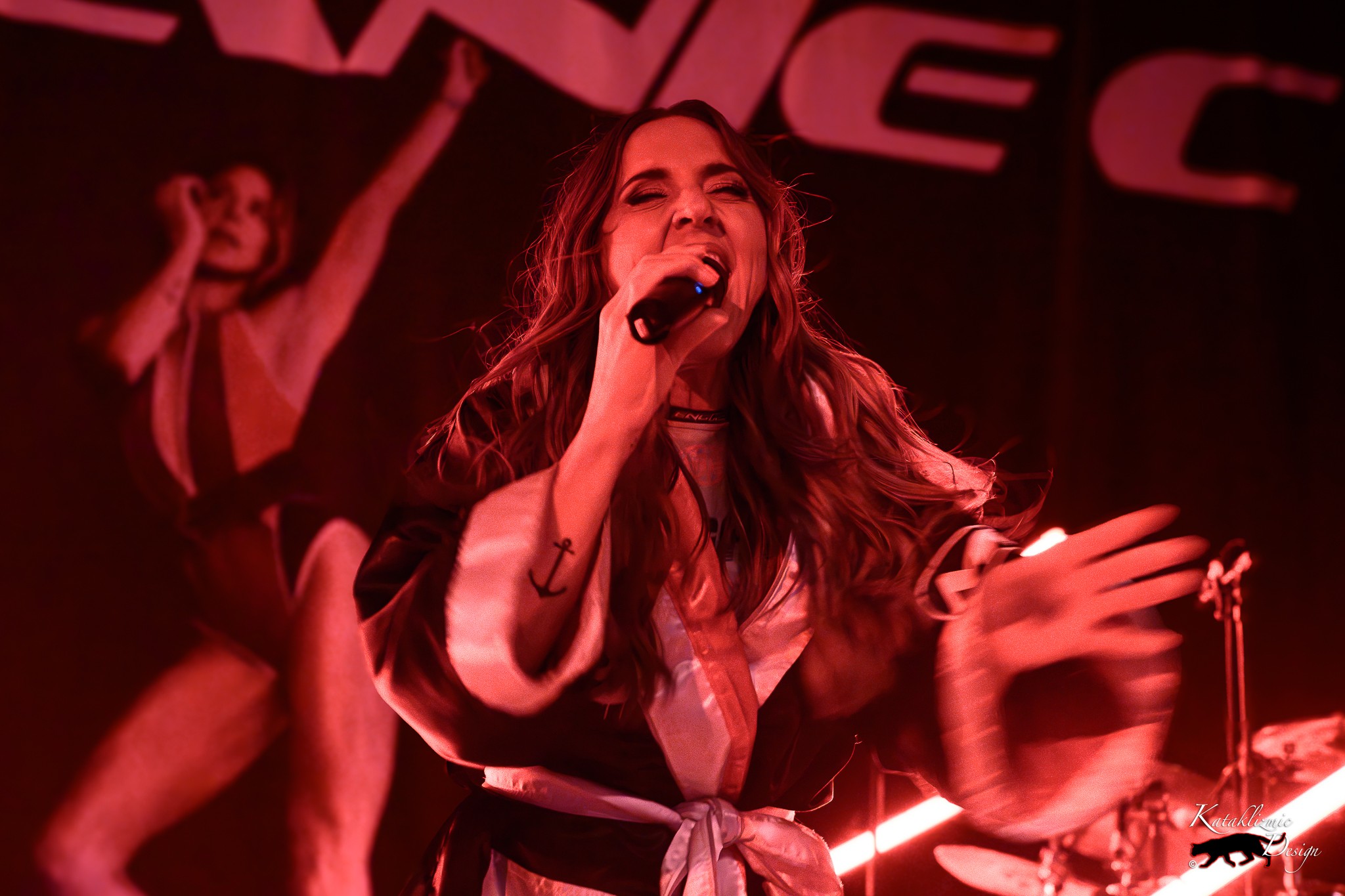 Melanie C singing into a microphone with one hand raised during a live performance under red stage lighting