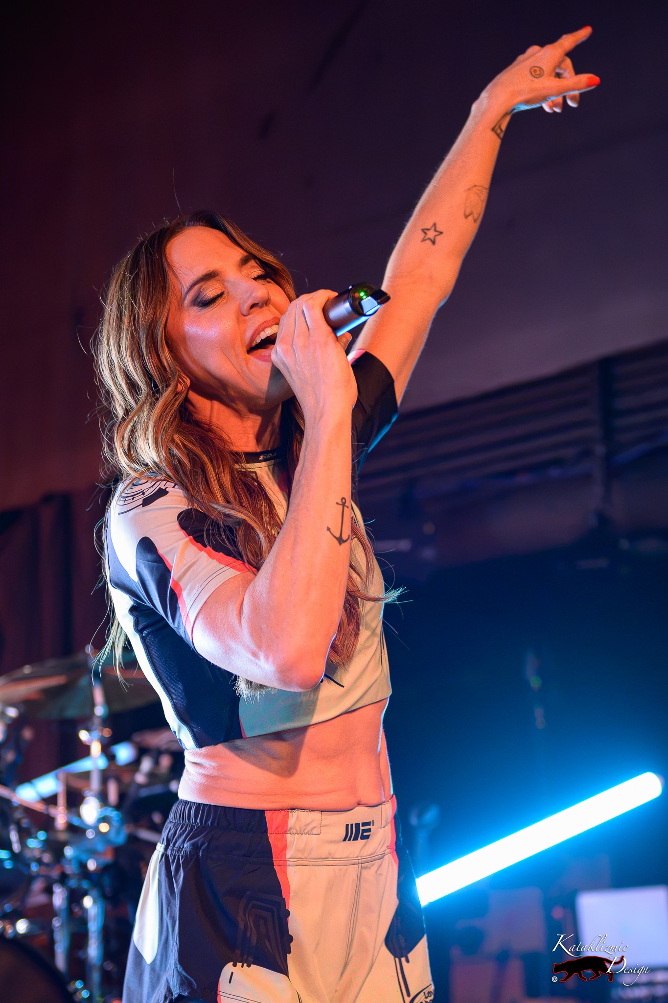 Melanie C singing into a microphone with one arm raised during a live performance under colorful stage lights