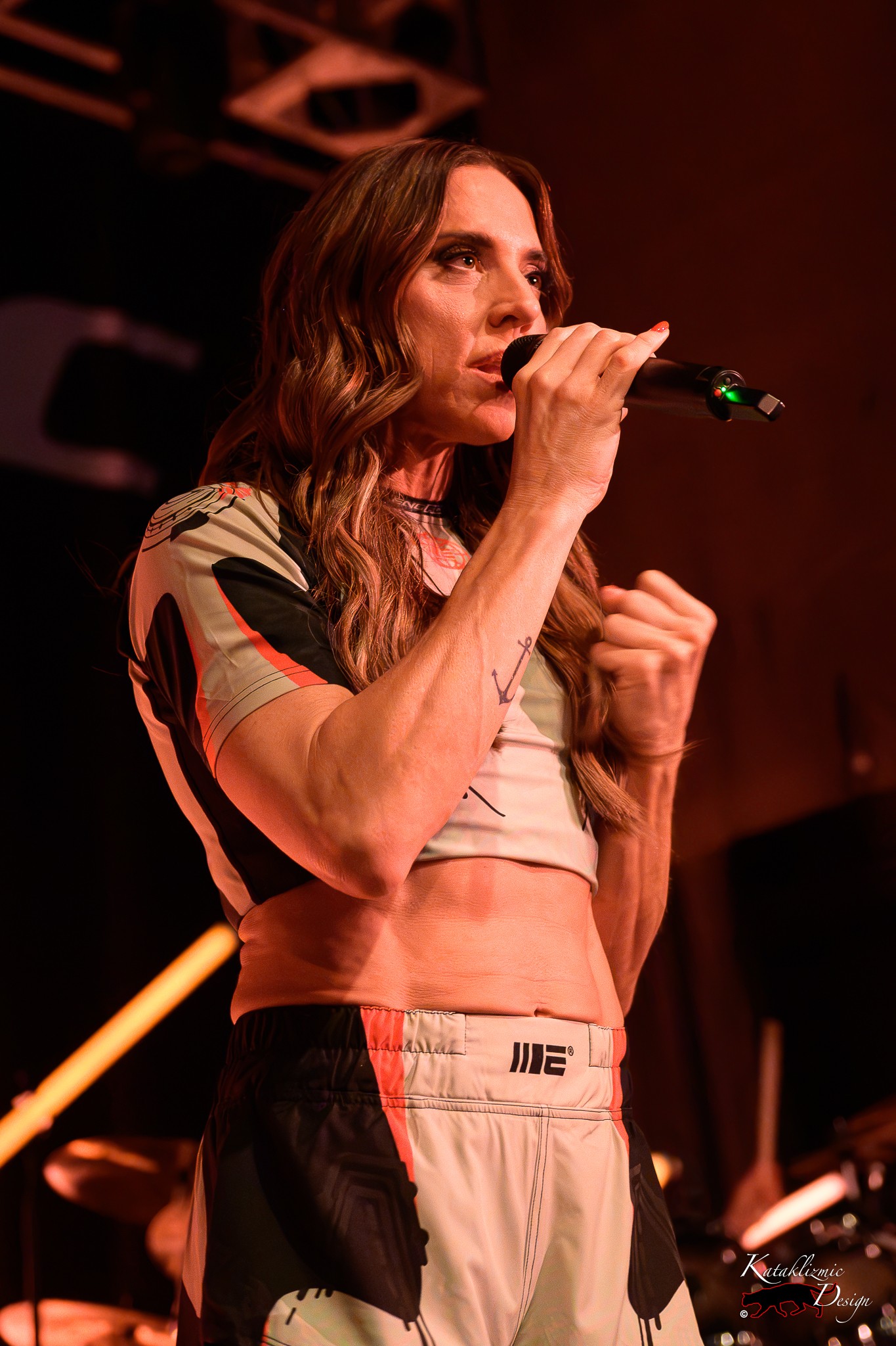 Melanie C sings into a handheld microphone under warm orange stage lights.