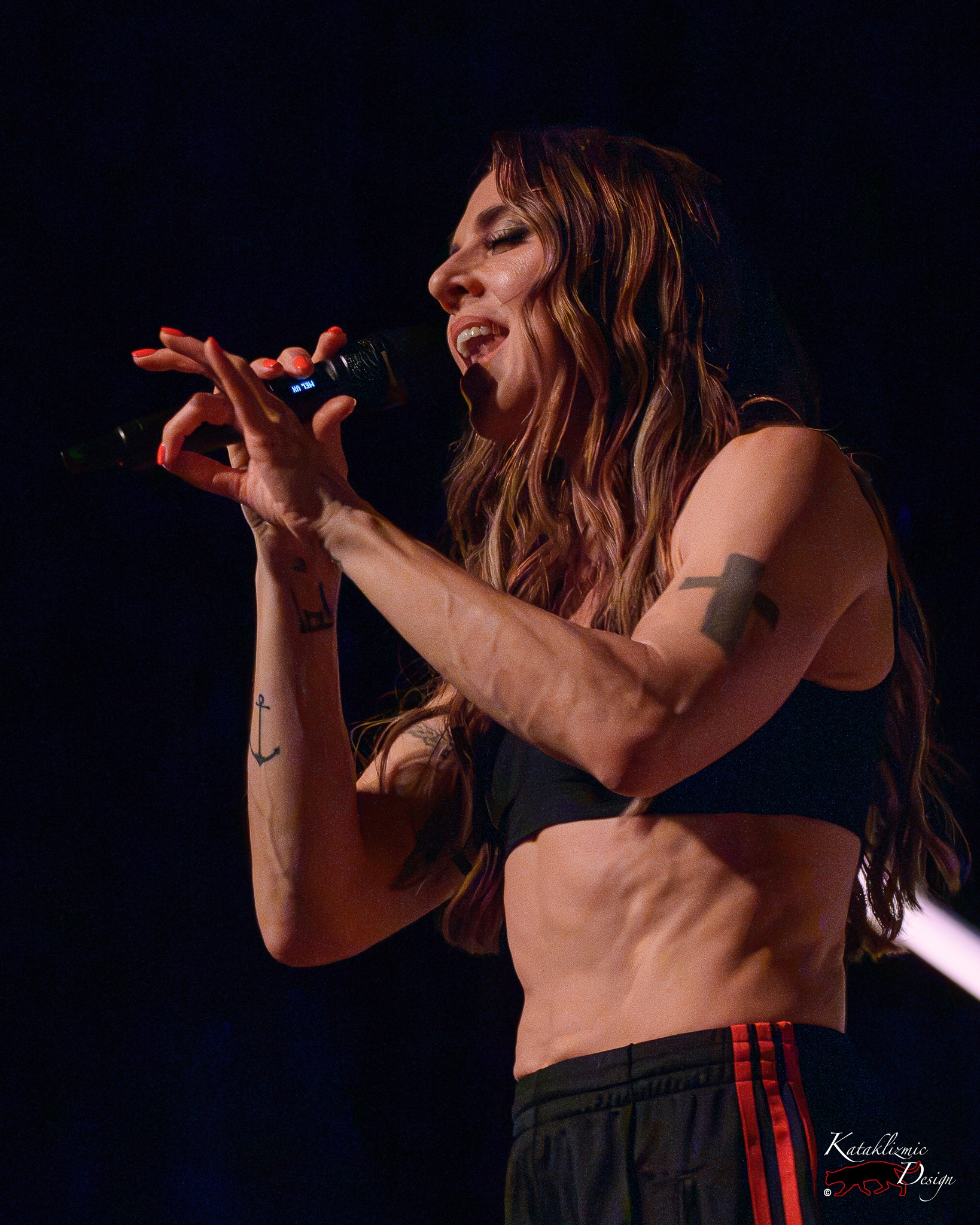 Melanie C sings into a microphone during a concert, her hands raised near the mic as dramatic lighting highlights her performance.