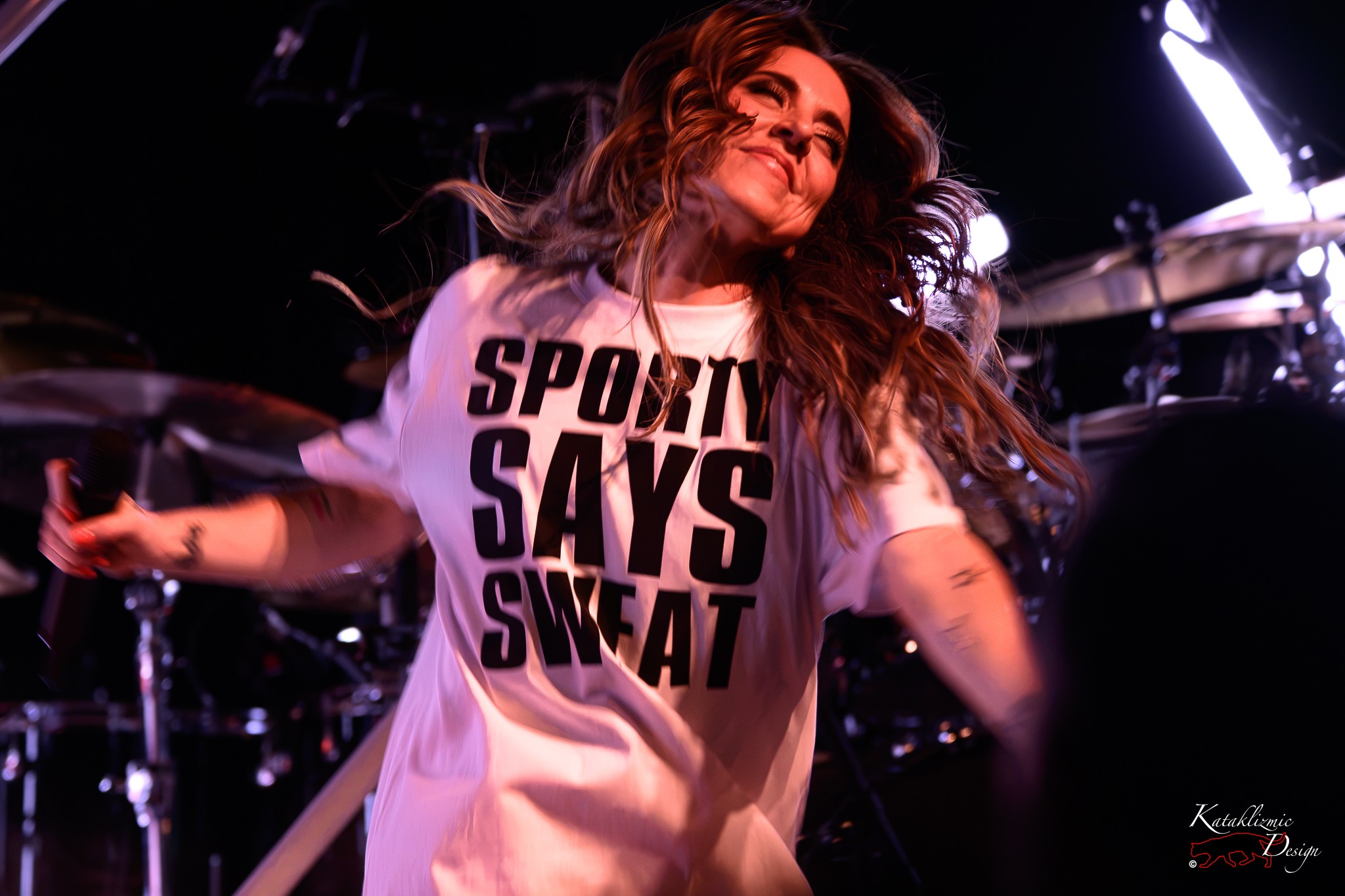 Melanie C performing live in a white “Sporty Says Sweat” shirt, smiling with her arms extended as her hair moves across her face under colorful stage lights.