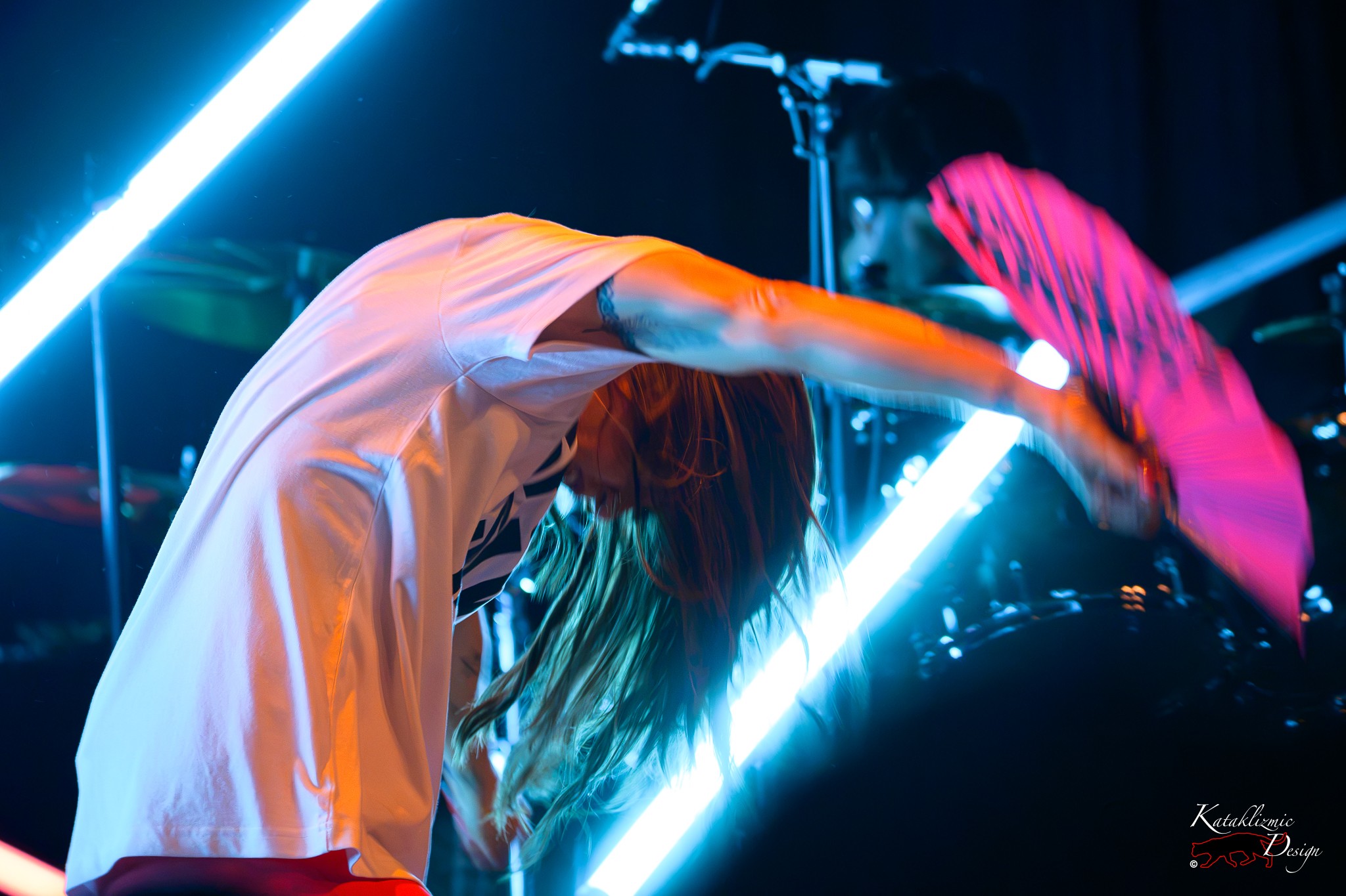 Melanie C bends forward with her hair in motion while extending a bright pink fan beneath blue and white stage lights at House of Blues San Diego.