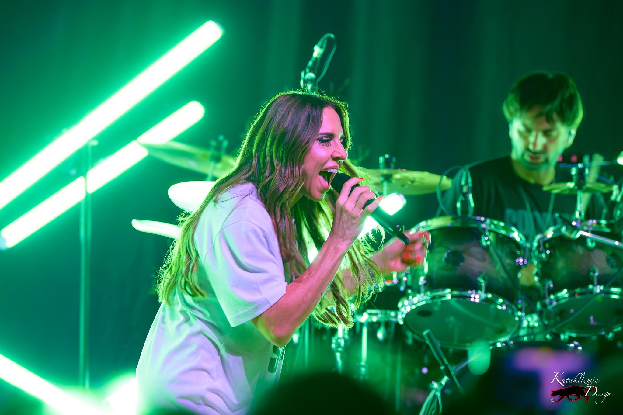Melanie C sings with a wide smile as her drummer performs behind her beneath vivid green stage lights at House of Blues San Diego.