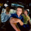 The Wldlfe band members pose inside a stripped-down vehicle, with one member seated in the foreground wearing a blue denim shirt and the others positioned behind him.