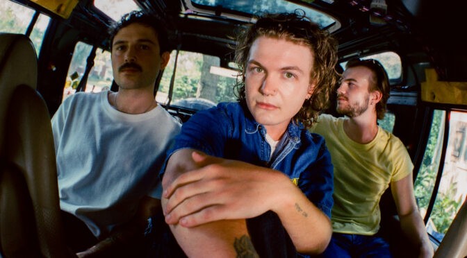The Wldlfe band members pose inside a stripped-down vehicle, with one member seated in the foreground wearing a blue denim shirt and the others positioned behind him.