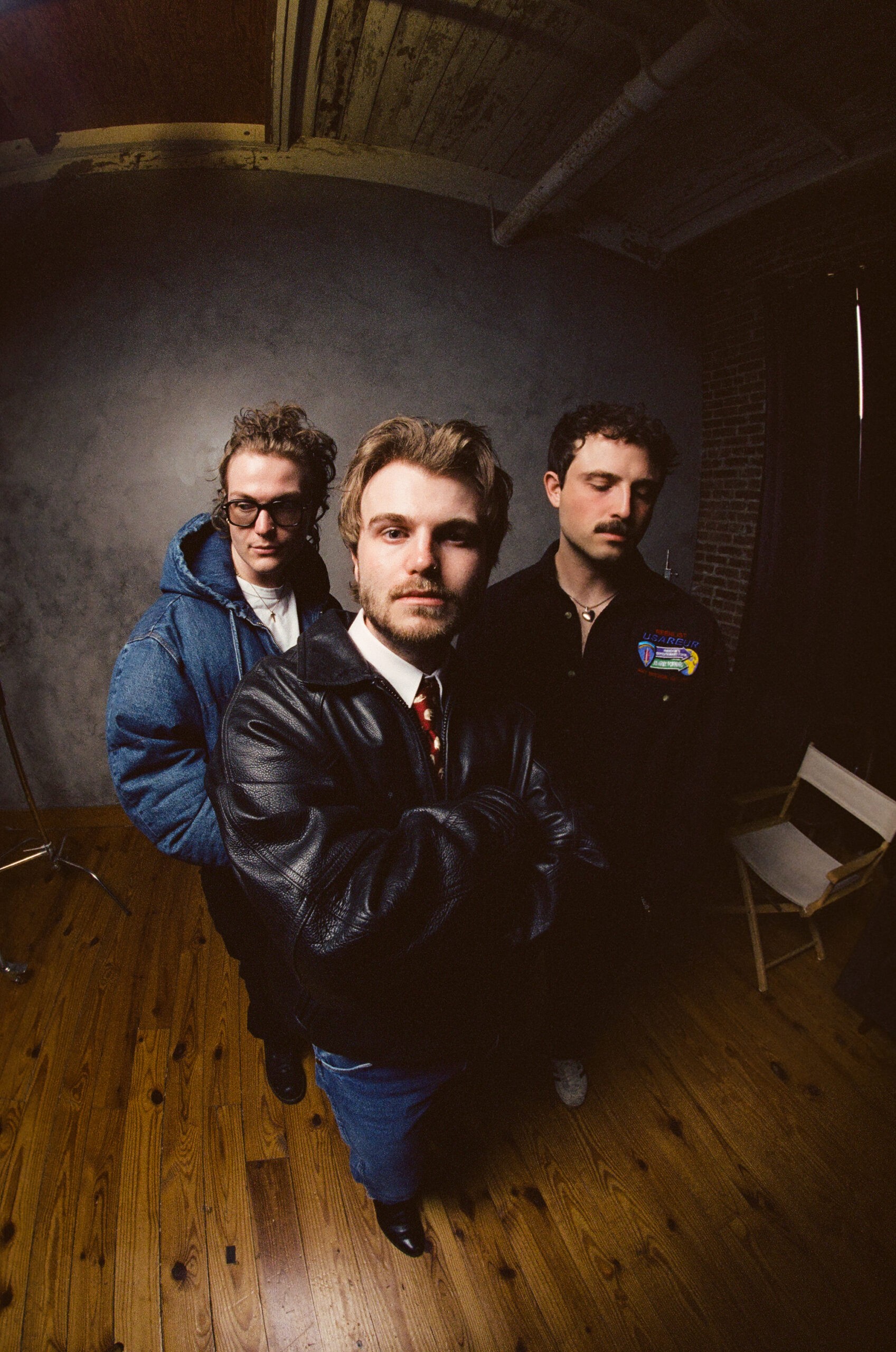 Indie-pop band The Wldlfe pose for a promotional portrait in a rustic room, with one member wearing a black leather jacket in the foreground and two others standing behind him.